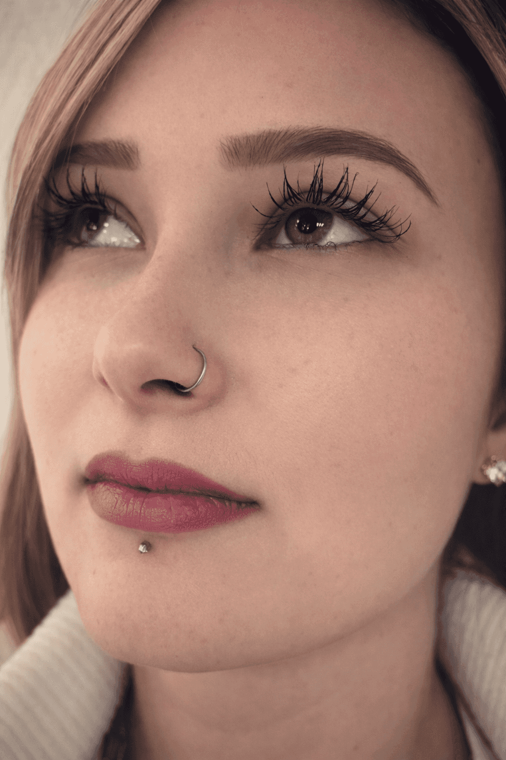 Femme avec un anneau au nez et un piercing à la lèvre regardant vers le haut, portant du mascara et un maquillage naturel, sur un fond neutre.