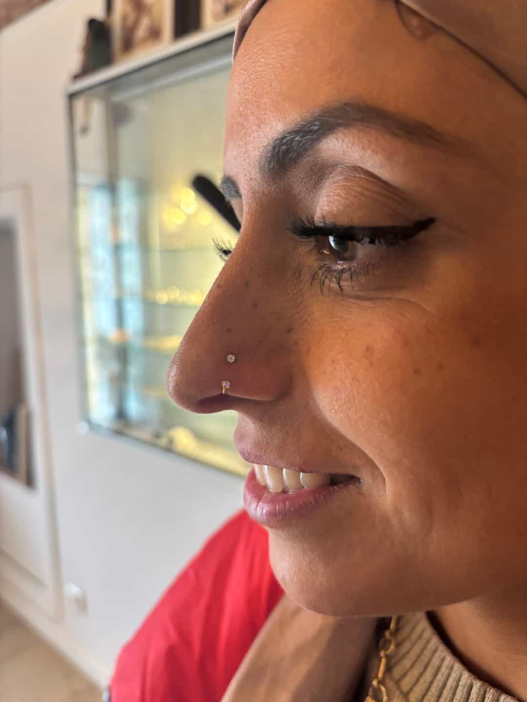 Femme souriante portant un hijab, avec un piercing au nez et un eye-liner ailé, se tenant dans une pièce avec une vitrine en verre en arrière-plan, réalisé par Nadia à Graphicaderme à Orange.